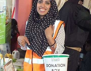 A picture of a smiling woman wearing a black head scarf with white polka dots, in an orange high visibility jack, carring a donation bucket
